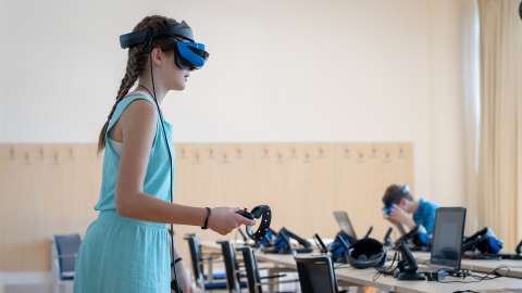 机の前に立って VR ヘッドセットを装着し、片手でコントローラを持ちながら VR ベースの授業に参加している学生