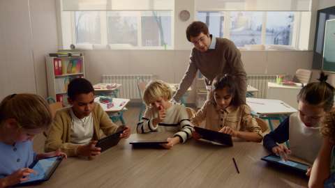 6 人の幼い生徒が教室のテーブルに座り、教師が 1 人の生徒の肩越しに作業の進捗を確認している