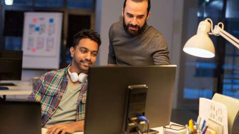 モダンでオープンな職場で、デスクトップ PC のモニターに表示された情報を共同でレビューする 2 人の同僚。1 人が座って微笑みながらキーボードに入力している一方で、もう 1 人は立って画面上の何かを指差している。